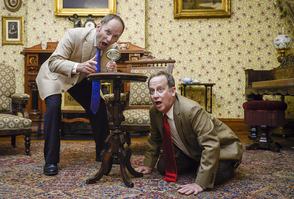 Photo of two murder and the macabre actors in the front parlor of the benjamin harrison mansion. one actor is crawling on the floor while the other leans over a table in dramatic fashion.