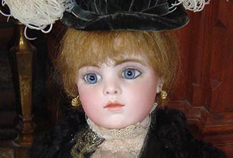 This close up photograph of the doll shows some finer details of the face, hat, and dress. Doll has Blonde hair and blue glass eyes. Her head shoulders, and forearm are bisque with upper arms & body thighs are kidskin leather. The lower legs and feet are celluloid. Tiffany mounted, fake pearl pierced earrings hand from her pierced ears. She is wearing a late nineteenth century style black dress with a feathered hat.
