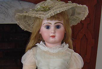 Photo of Baby Mckee's jumeau doll. The head is bisque and composition body has jointed arms and legs, blue glass eyes and long curled blond hair. The doll has pierced ears with imitation ruby earrings.