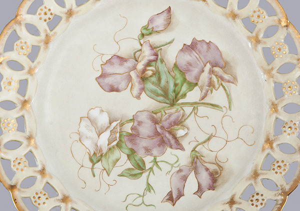 Photograph of plate with hand painted sweet peas. Edge is a cutout lace pattern with gold paint. Called a ribbon plate because ribbon could be put through border to hand on wall.
