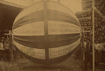 Photograph (sepia tone) mounted on heavy board. Images shows 1888 Campaign Ball with D. E. Brockett (designer and builder) standing to the left with other men behind him and to the right side of the ball. There is also a stack of lumber to the back on the right and a shed type building to the left. Below image on board is the following information: 