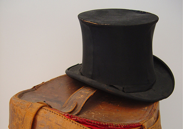 Benjamin Harrison's silk top hat on top of a leather carrying case. Black satin or silk hat. the hat is 6 inches tall with a slight bowed flare toward the middle the stack; seams on both sides; a band to cover the joining of stack and brim is missing except on the right front; a partial bow is placed on the right side; stitches are showing where band would have been.