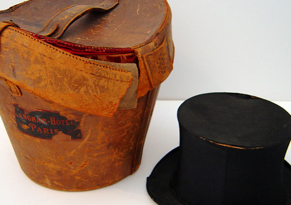 Leather carrying case for Benjamin Harrison's top hat. Case: Leather shows signs of red rot. Straps broken, rim of lid coming detached from both sides, and general marks and scuffs on leather. Harrison's black silken top hat lays next to the case for scale and reference.
