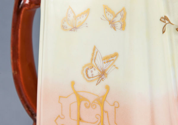 close up photo of the chocolate pot. shows intricate gold painting of three butterflies floating above ornately painted initials, E.H. for ethel hurty.