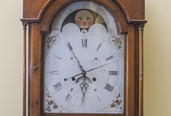 Tall case or grandfather clock made by George Woltz in Hagerstown, Maryland between 1800 and 1812. Clock has moon dial at top and a date dial below the center of the hands, and strikes on the hour. Cherry cabinet with front legs that curve out . There is a spindle on either side of the face. Rounded center at the top with rosette design. Top corners come up to pointed spindles. Clock face is a half circle at top with the dates above the moon dial, Roman numerals and painted floral designs in four corners, and glass door.