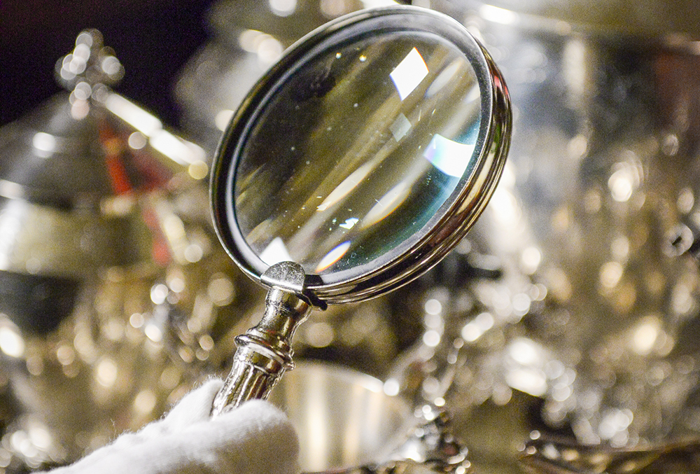 Photograph of a magnifying glass being held by a gloved hand in front of a blurry background.