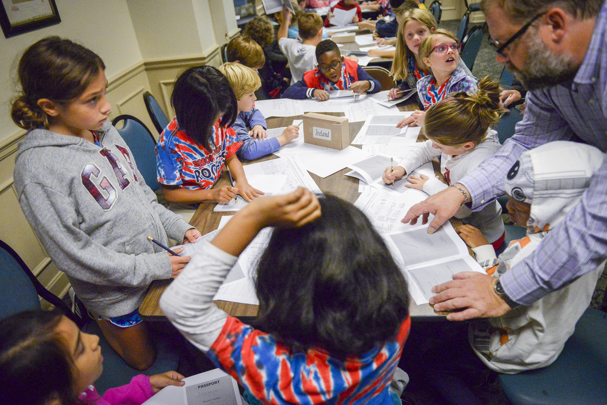 Photograph of staff member Roger Hardig helping young students at the benjamin harrison site. The table is cluttered with loose papers and some students are writing.