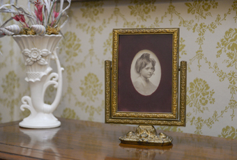Framed photo of Elizabeth Harrison. Oval photograph window in maroon velvet mat. Gold painted wooden easel type frame with fancy metal base holding wood frame that tilts or swivels up and down.