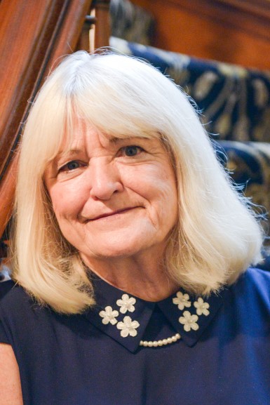 Photograph of Hazel Gillaspy. She is an elderly women wit platinum blonde hair. She wears a blue blouse with a collar that is embroidered with a floral design with a pearl necklace underneath. She has light colored eyes.