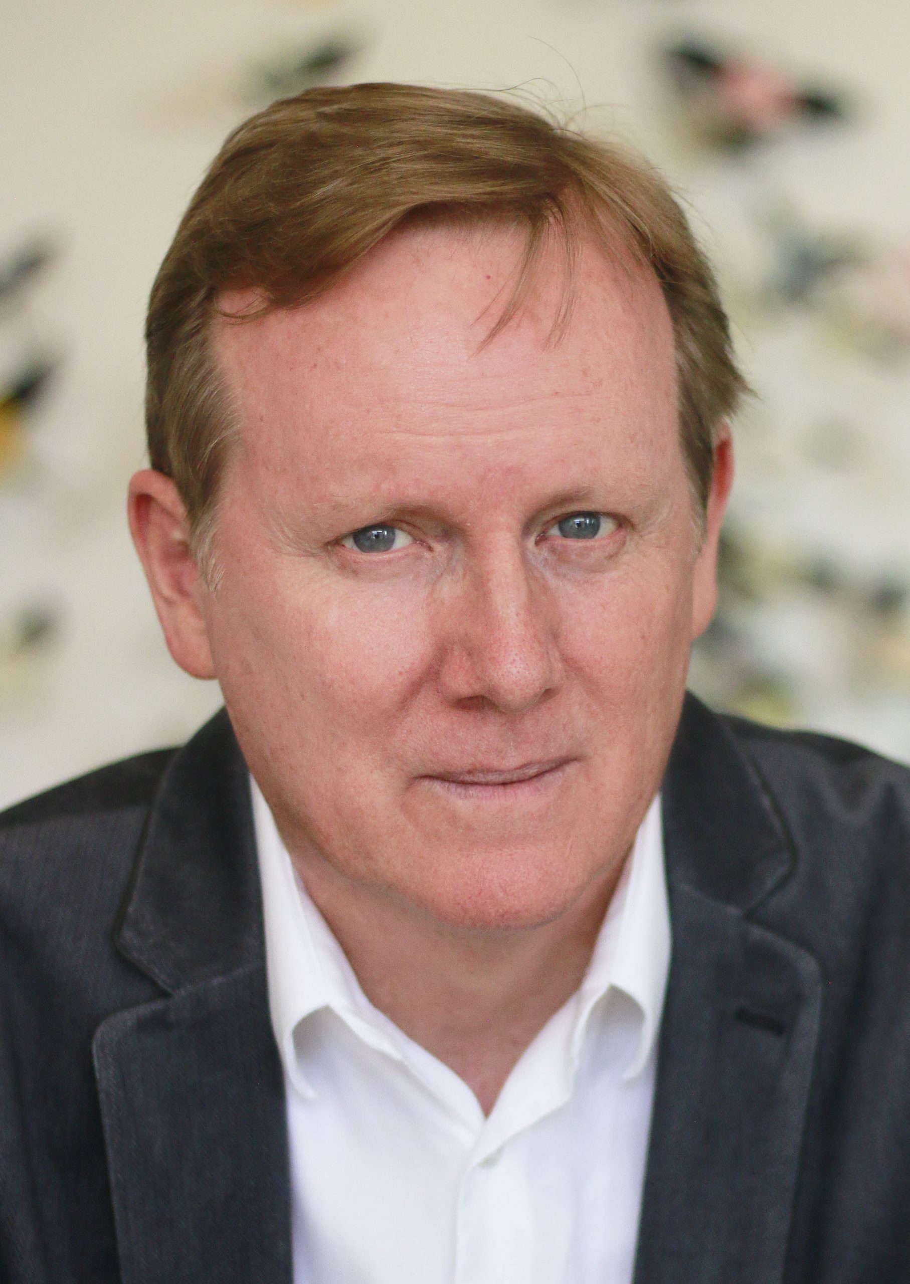 Portrait of Dave Surina. A middle aged man with light brown hair and green eyes. He wears a black jacket with white dress shirt, in front of an out of focus floral background.