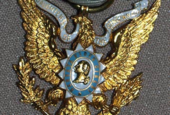 Daughter of the Cincinnati badge. Predominantly white ribbon with blue edging holding a golden eagle. An emblem in the center of the eagle contains a golden bust of a male figure and is encircled by the text 