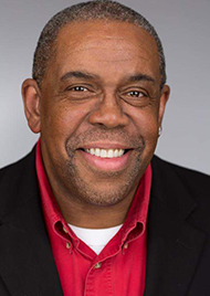 Portrait of Dennis Jones. He is middle aged and has short dark hair and brown eyes. He wears a white undershirt, red dress shirt, and black jacket, and smiles at the camera.