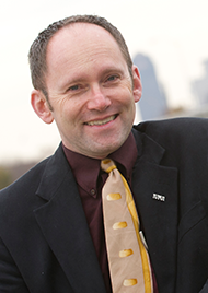 Portrait of Stephan Viehweg. A middle aged man, he has short brown hair and no facial hair. He wears a crimson dress shirt, black jacket, and patterned yellow and beige tie.