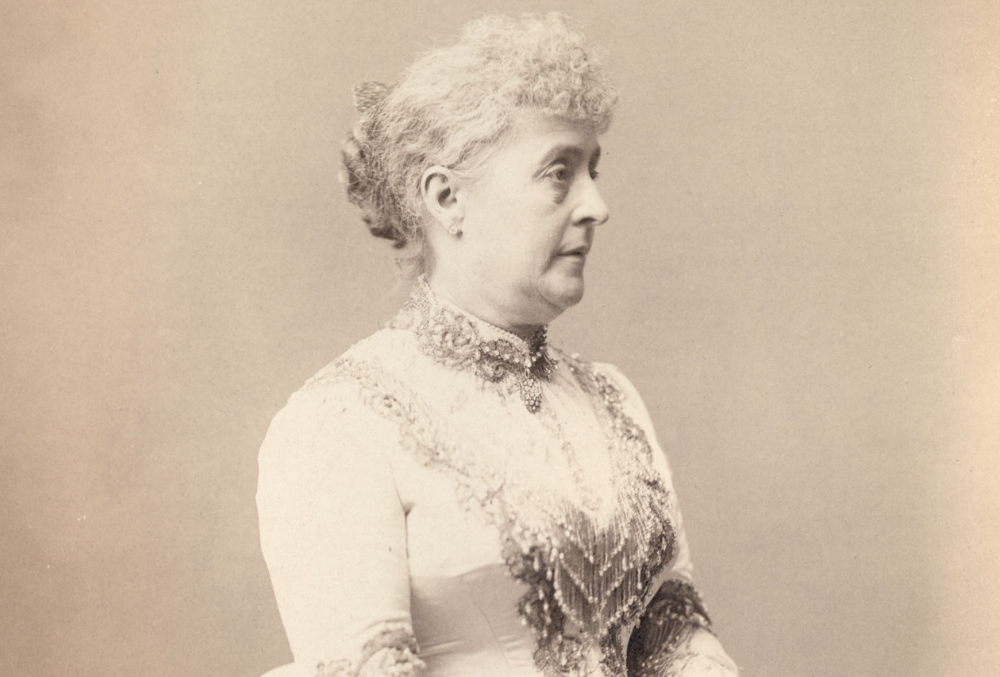 Sepia-toned photographic image of Caroline Harrison in her inaugural dress which has a large bustle and beading on the bodice and around the neck. Caroline is wearing gloves and holding a fan; her hair is pulled back from her face. She is standing in front of a large bookcase and is resting her hands in the back of a chair, while holding a fan and kerchief.