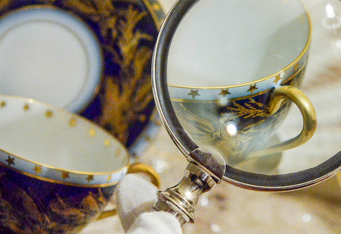A photograph of an intricate coffee cup, complete with gold lining, gold handle, and stars along the mouth of the cup. There is a magnifying glass being used to enhance the detail.