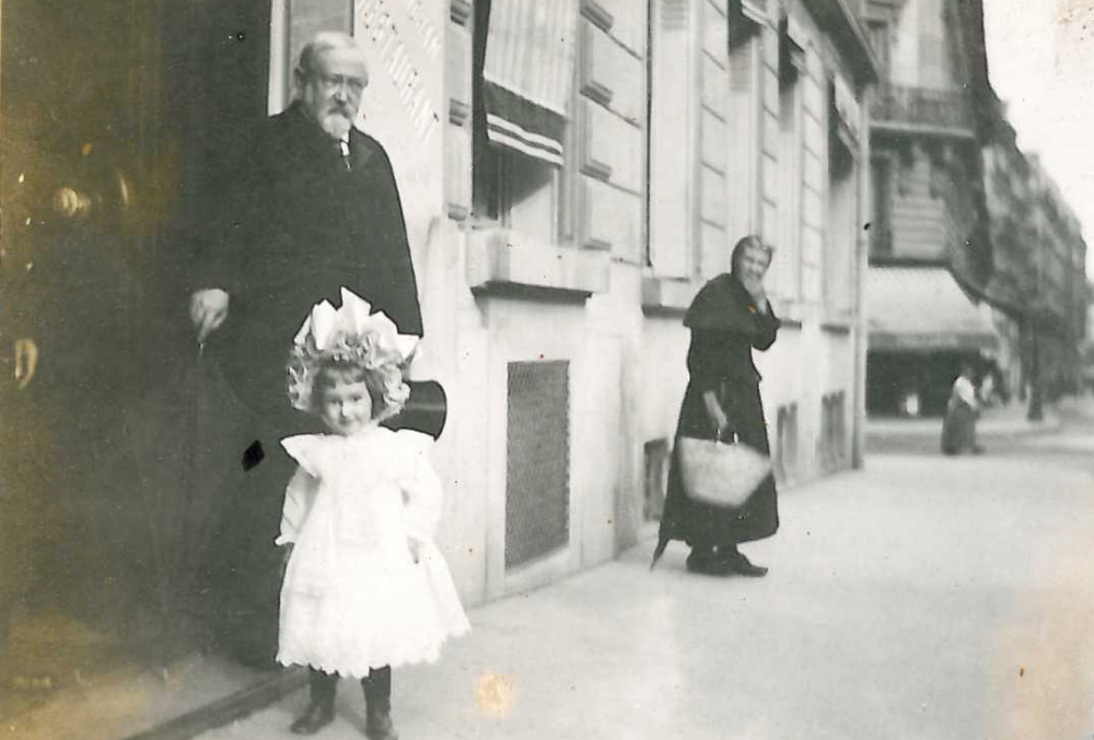 Photograph of Benjamin Harrison and his daughter, Elizabeth Harrison Walker, on a street in Paris. Benjamin Harrison steps out of a building with his cane in his right hand, and his top hat in his left. Elizabeth wears a frilly white dress and a frilly white hat. Slightly out of focus in the background is a woman carrying a tote bag with an expression of surprise at seeing Benjamin Harrison.