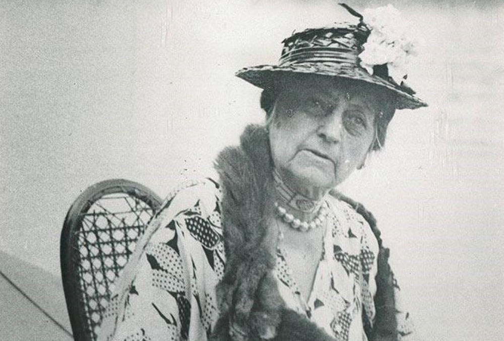 Photo of Mary Dimmick Harrison, pictured seated in a wooden woven chair, wearing a patterned blouse, fox pelt scarf, pearl necklace, and woven hat with a corsage.